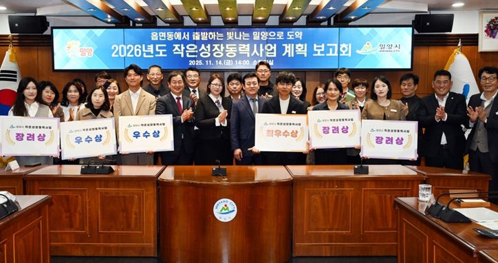 밀양시청 소회의실에서 열린 2026년도 작은성장동력사업 계획 보고회에서, 평가 결과 선정된 읍·면·동 직원들이 사업 선정 표찰을 들고 단체 사진을 촬영하고 있다.